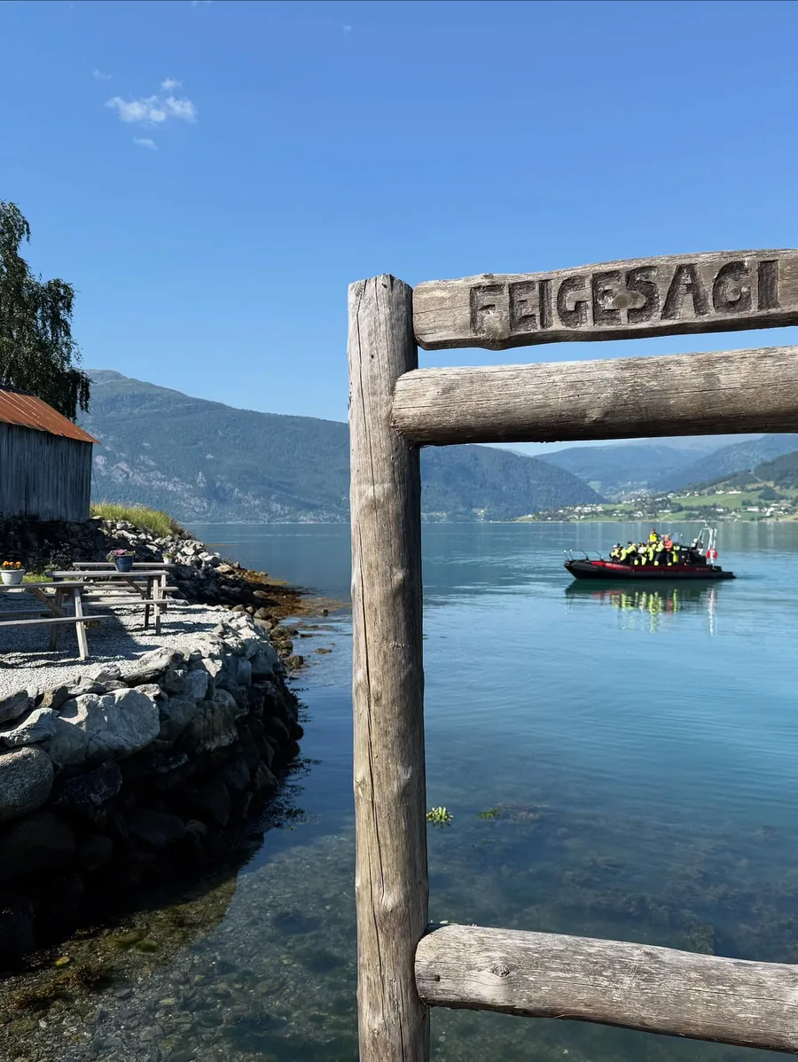 The carved Feigesagi sign on a timber frame, with Lustrafjorden and a boat behind it