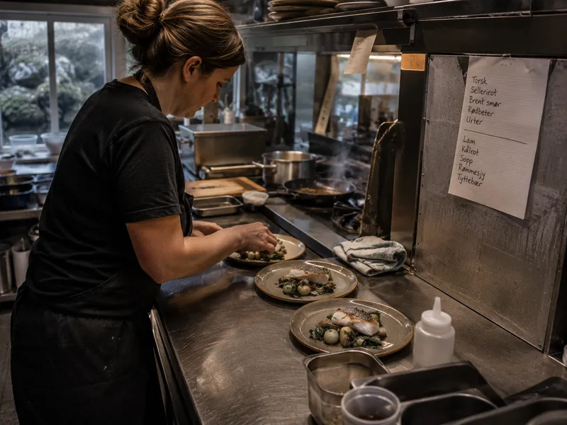 A cook plating fish at the pass in a small kitchen, handwritten menu on the wall