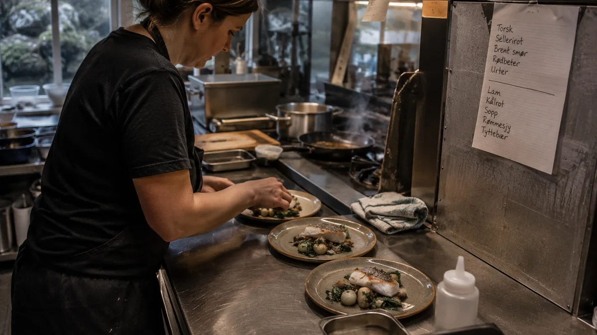 A cook plating fish at the pass in a small kitchen, handwritten menu on the wall
