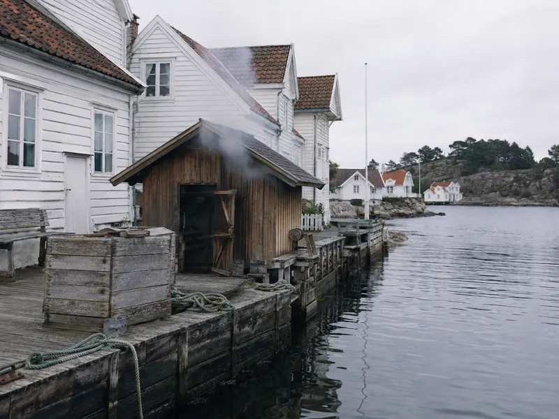 A timber smokehouse on the quay with smoke rising, white houses and grey water behind