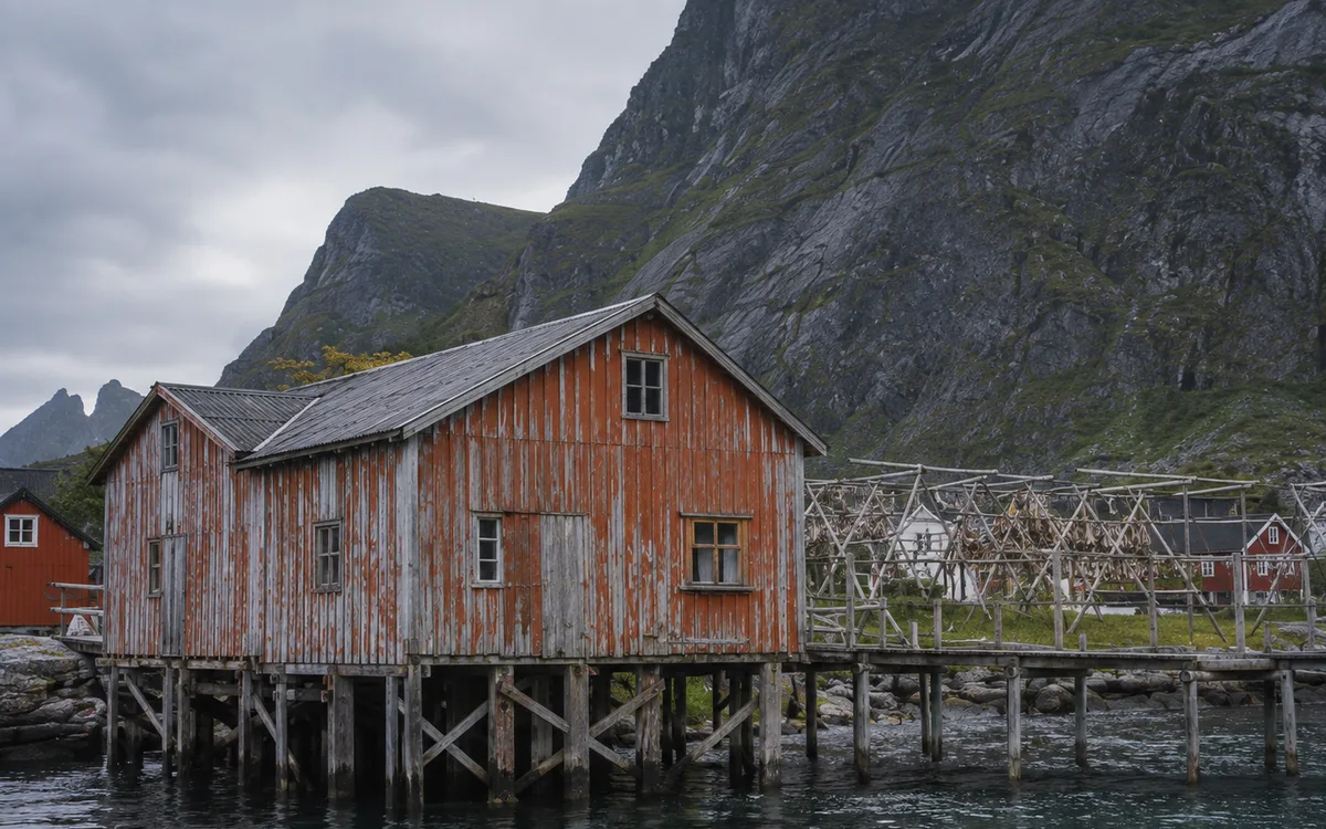 Weathered timber fish warehouse on piles over the water in Henningsvær, drying racks behind