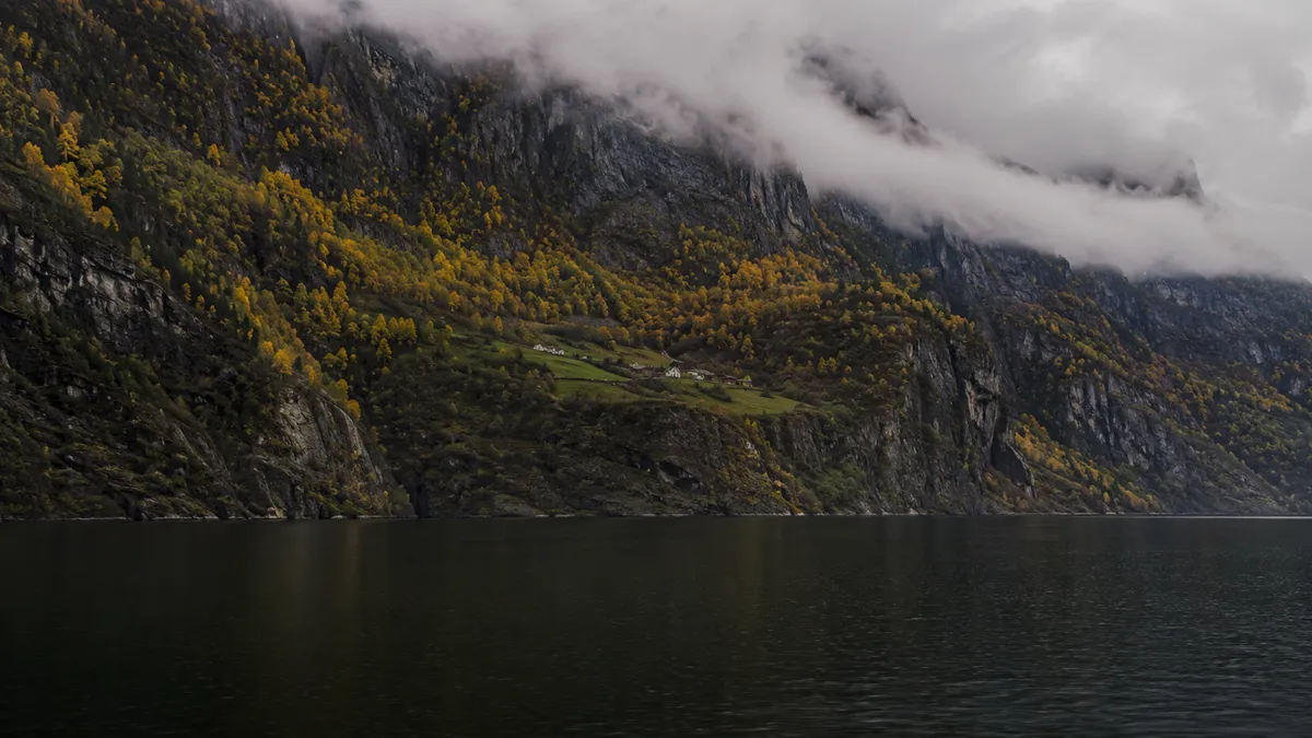 Gårdsbruk på en grønn hylle høyt over Sognefjorden, med lav sky langs fjellveggen
