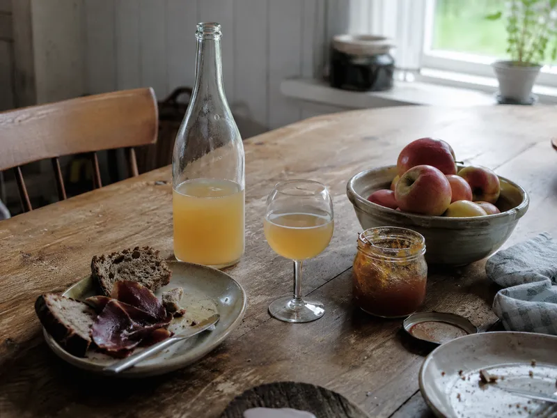 A bottle of cloudy cider, apple preserve, bread and cured meat on a worn kitchen table