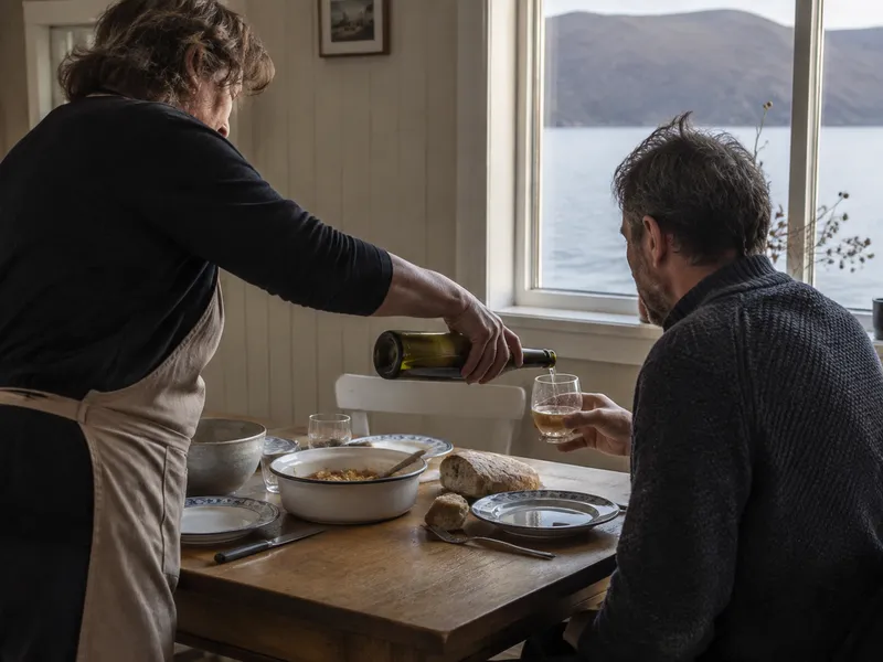 The host pours cider for a guest at a plain table by the window