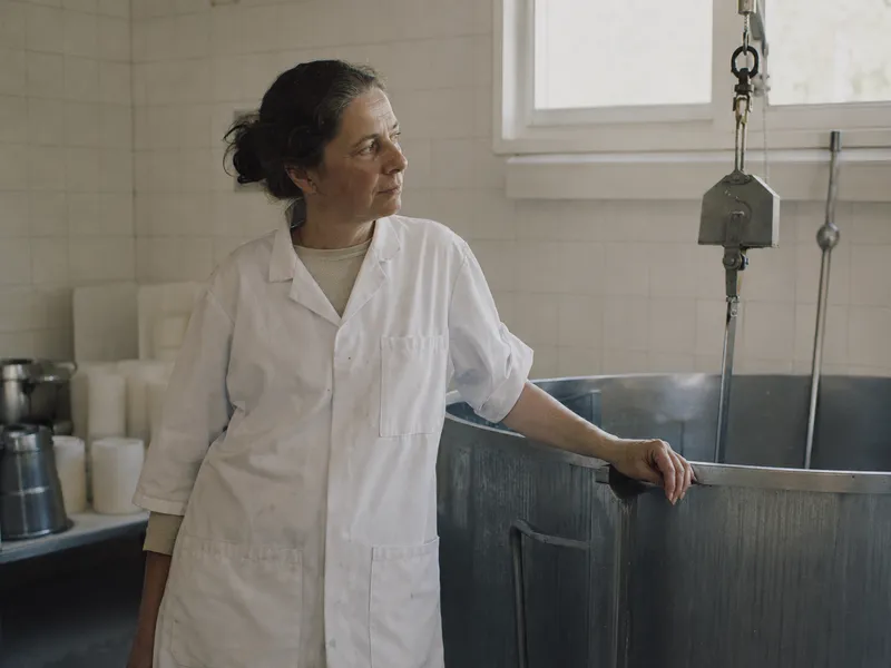 A cheesemaker in a white coat beside the vat, looking away from the camera